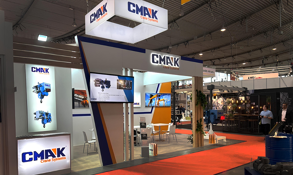 The modern exhibition booth of CMAK Crane Company at an international industrial trade show, displaying digital product presentations and wire rope hoist systems.