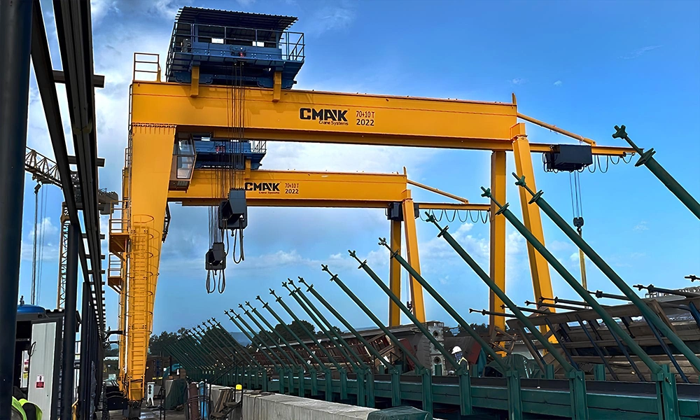Two heavy-lift 70-ton capacity double-girder gantry cranes CMAK with integrated operator cabins and custom weather-roof open winches installed at an outdoor industrial infrastructure shipyard.