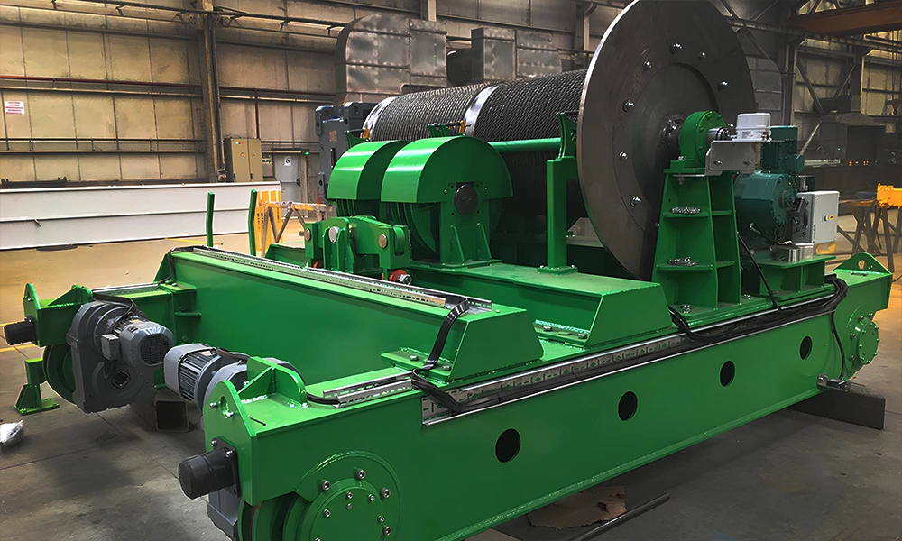 A high-capacity green open winch trolley assembly from the special cranes CMAK line, featuring a massive wire rope drum, heavy-duty industrial gearboxes, and multi-wheel travel end carriages on a factory assembly floor.