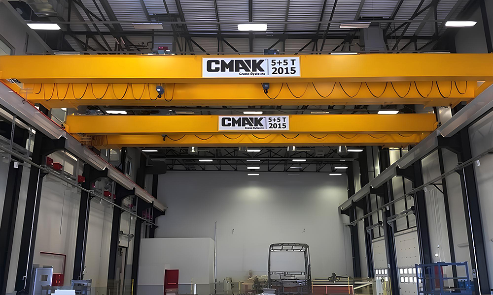 Two yellow CMAK double-girder bridge cranes installed inside a modern industrial warehouse and production facility.