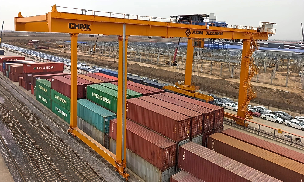 Large yellow double-girder rail-mounted container handling crane from the gantry cranes CMAK line operating at the ADM Jizzakh facility, lifting and stacking shipping containers next to railway tracks.