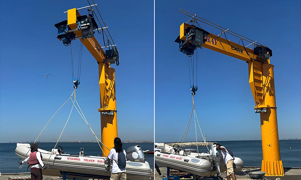 A yellow pillar-mounted workstation crane from the jib cranes CMAK series installed at a coastal marina, lifting an inflatable boat with an outboard motor out of the water.