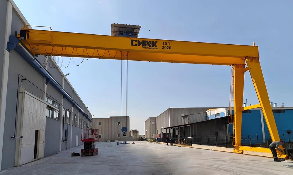 An outdoor 10-ton yellow semi-gantry crane from the gantry cranes CMAK product line, configured with one side running on an elevated building runway rail and the other side on a ground track.
