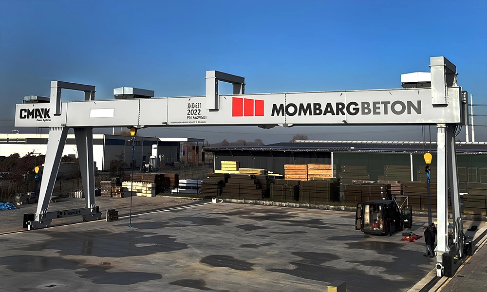 A heavy-duty outdoor gray double-girder rail-mounted crane operating as part of the gantry cranes CMAK product line at the Mombarg Beton precast concrete facility under a clear blue sky.